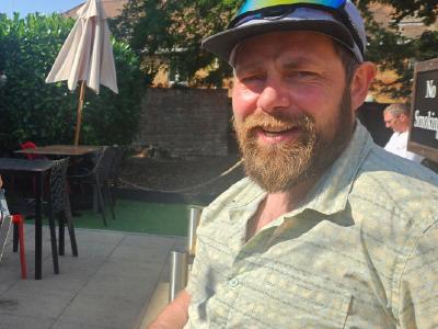 Two cyclists resting at outdoor pub patio; left cyclist wears blue cap, sunglasses and black kit with red gloves; right cyclist has beard, cap with sunglasses on brim; tables and umbrellas behind them.