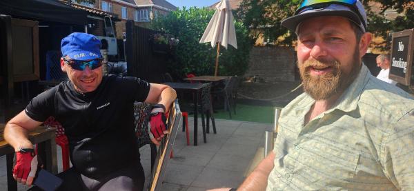 Two cyclists resting at outdoor pub patio; left cyclist wears blue cap, sunglasses and black kit with red gloves; right cyclist has beard, cap with sunglasses on brim; tables and umbrellas behind them.