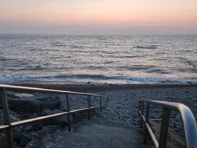 Pebble beach at dawn with metal handrails leading down to gentle waves, wooden groynes along shoreline, soft pink and orange sky gradient over calm sea.