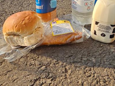 Cycling provisions on gravel ground including bread roll, baked potato, canned beans, large water bottle and milk carton, red hatchback car and houses in background.