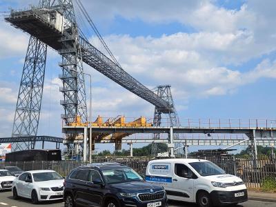 Industrial transporter bridge with steel lattice towers and suspended gondola structure spanning overhead, cars parked in foreground under blue cloudy sky.