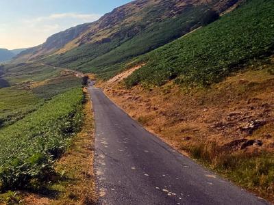 Single-track lane winding through wide green valley between steep hillsides with stream running along left edge under bright late afternoon sun.