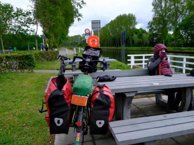 Touring bike with panniers parked by a canal‑side picnic table where a person sits.