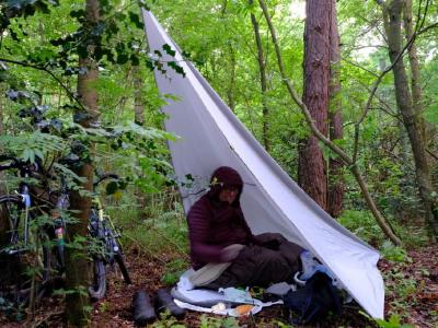 Person in bivy bag under a white tarp shelter with bikes in dense woodland.