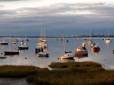 lowerhalstow.jpg A harbor scene at dusk with numerous small boats and yachts moored in calm water, featuring a cloudy sky with warm evening light and grassy salt marshes in the foreground.
