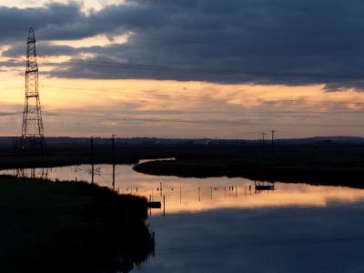 reflections.jpg A dramatic sunset landscape showing a winding waterway reflecting cloudy skies, with an electrical transmission tower silhouetted against the golden and blue evening sky.