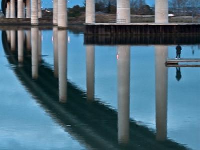 sheppeycrossing.jpg A modern concrete bridge with multiple cylindrical support pillars crossing over calm water, creating perfect reflections of the structure and overcast sky in the still water below. The bridge appears to curve slightly and has a person visible on a small platform or dock near the water's edge.