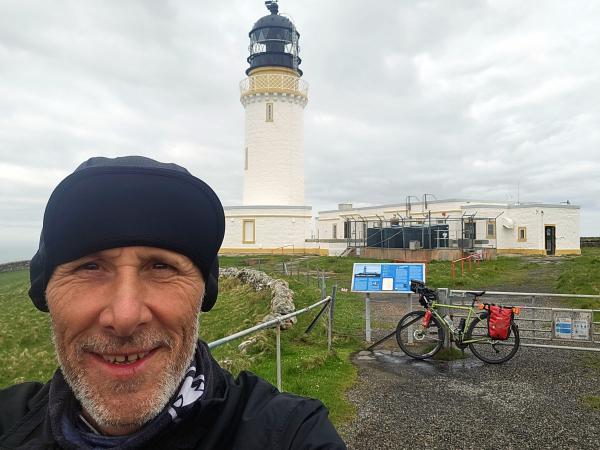 Close-up of a green touring bicycle with red panniers parked by a metal gate near a white lighthouse with a black lantern room and surrounding outbuildings, set in grassy terrain with a cloudy sky overhead.