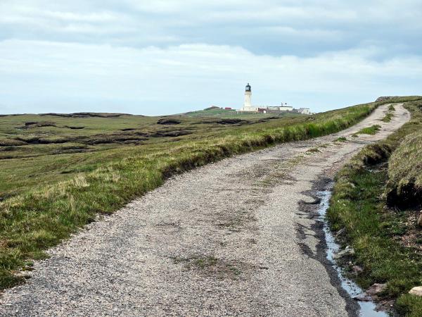A rugged track leads toward a distant white lighthouse and its outbuildings, set on rolling green hills with a cloudy sky above.