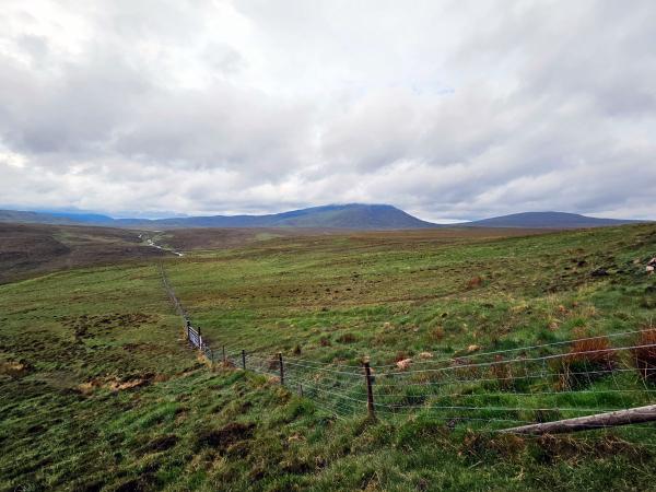 A panoramic view across wide, open moorland with gently sloping green hills. A wire fence runs diagonally into the distance, and a mountain peak is visible under dramatic cloud cover.