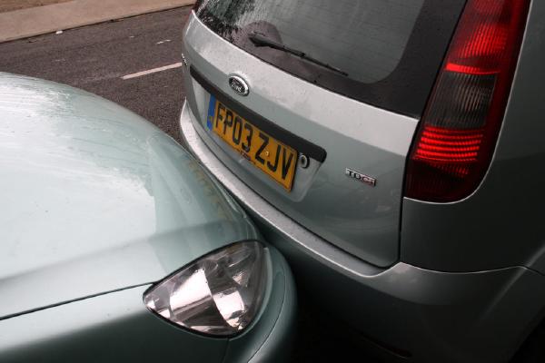 A silver car is parked with its rear bumper pressed up against the front bumper of another light green car on the street, showing no visible damage. Both vehicles are wet from rain, with the silver car&rsquo;s number plate &ldquo;FP03 ZJV&rdquo; clearly visible.