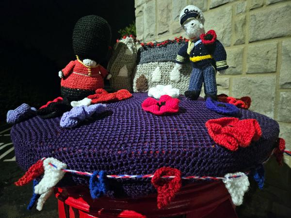 A handcrafted Remembrance Day tribute featuring knitted figures and elements arranged on a purple knitted postbox topper. The scene includes a British Queen&rsquo;s Guard soldier in red uniform with black bearskin hat on the left, a miniature knitted cenotaph monument in the center draped with a red poppy garland, and a naval or air force officer figure in navy blue uniform on the right. Multiple red knitted poppies are scattered across the purple surface, along with small red, white, and blue decorative elements. The display sits on a red postbox against a stone wall background.