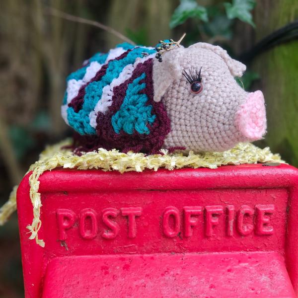Crocheted multicoloured pig postbox topper with long eyelashes sitting on a red “Post Office” box in Barham, Kent.