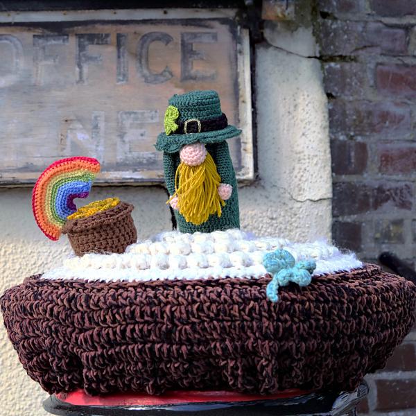 Crocheted post box topper in Shorne Village, Kent showing a leprechaun with yellow beard, pot of gold and rainbow on a textured white and brown base.