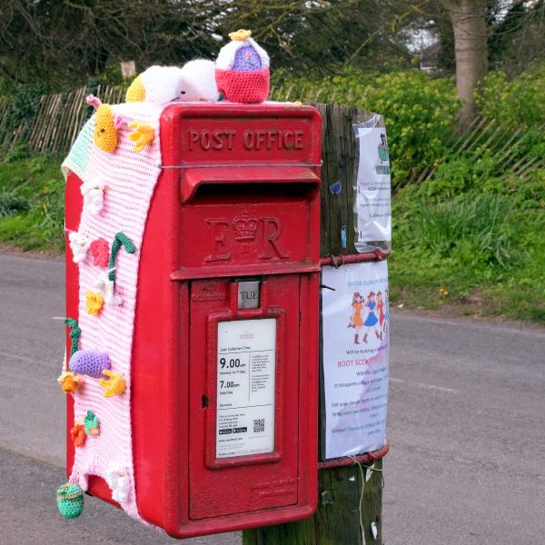 Red village postbox decorated with crocheted Easter designs beside a roadside.