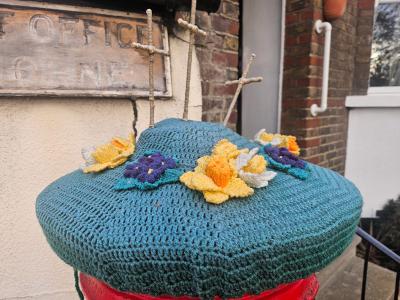 Hand-crocheted Easter post box topper in teal featuring three beige crocheted crucifixes, decorated with spring daffodils in yellow and white and purple crocuses, displayed on a red Royal Mail post box in Shorne Village