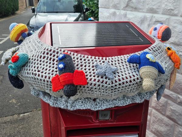 A red postbox of the future with a solar panel on top is wrapped in a chunky crocheted grey topper decorated with knitted space‑themed figures, including rockets, planets, stars and a tiny orange astronaut.