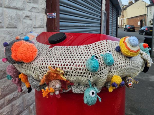 Rear view of a red postbox covered by a grey crochet topper showing woolly planets, bug‑eyed aliens and pom‑pom details clustered around the rim.