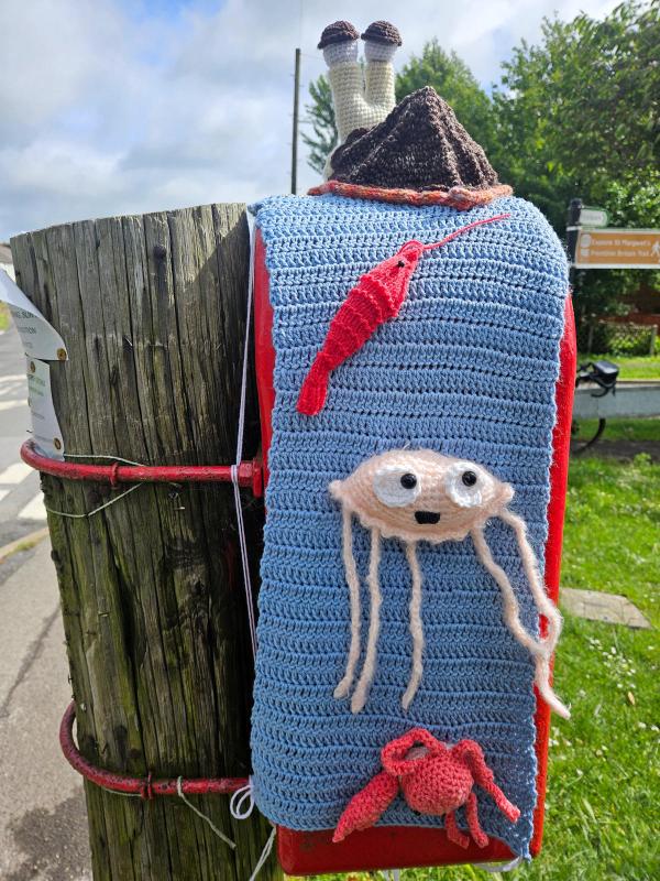 Red Royal Mail post box decorated with yarn bombing. A pale yellow crochet trim covers the &lsquo;Post Office&rsquo; lettering on the box door. A large white crochet snail with tall stalk eyes and brown eyebrows perches on top. Residential street visible in background.