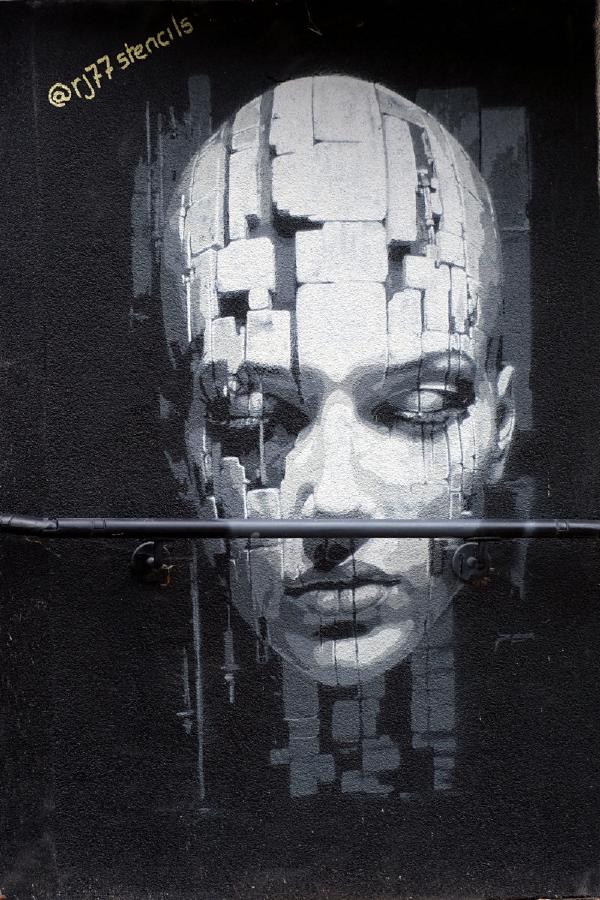 Black-and-white mural of a solemn human face on a dark wall, composed of pixel-like blocks, with a horizontal metal pipe crossing at mouth level and @rj77stencils stenciled in yellow at top left.