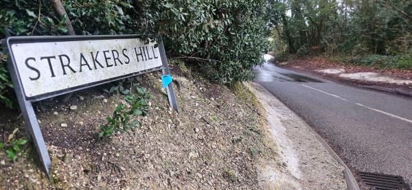 Street sign reading “STRAKERS HILL” on a bank beside a quiet country road with wet tarmac and trees ahead.
