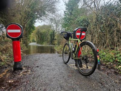 Narrow tree-lined lane heavily flooded with brown rainwater, with a muddy verge and wooden handrail on the left.