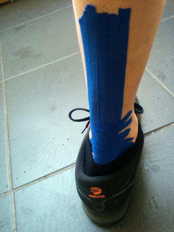 A close-up view of a cyclist&rsquo;s lower leg and foot showing bright blue kinesiology tape applied from ankle to mid-calf, wearing a black Adidas training shoe on tiled flooring.